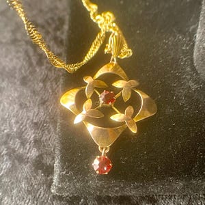 May include: A gold-toned pendant necklace featuring a four-leaf clover design with a central red gemstone and four smaller gemstones. The pendant hangs from a delicate, twisted gold chain. The necklace is displayed against a dark background.