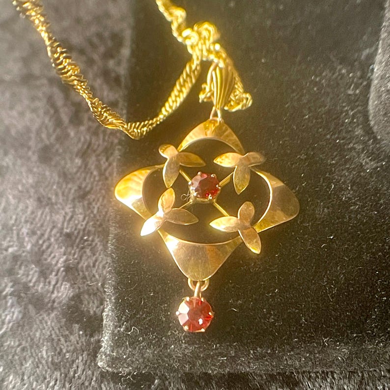 May include: A gold-toned pendant necklace featuring a four-leaf clover design with a central red gemstone and four smaller gemstones. The pendant hangs from a delicate, twisted gold chain. The necklace is displayed against a dark background.