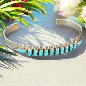 Op de afbeelding: Een zilveren manchetarmband met turkooizen stenen. De armband heeft een rij turkooizen stenen gezet in zilver, met een gedetailleerd ontwerp langs de randen. De turkooizen stenen zijn helder blauwgroen.