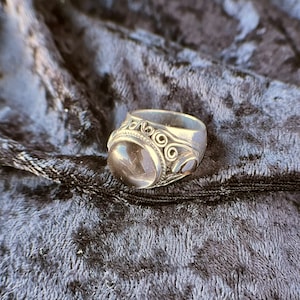 May include: A silver ring with a large, clear, rounded stone as the centerpiece. The ring has decorative swirls and two smaller stones on the sides. The ring is set against a dark, textured background.