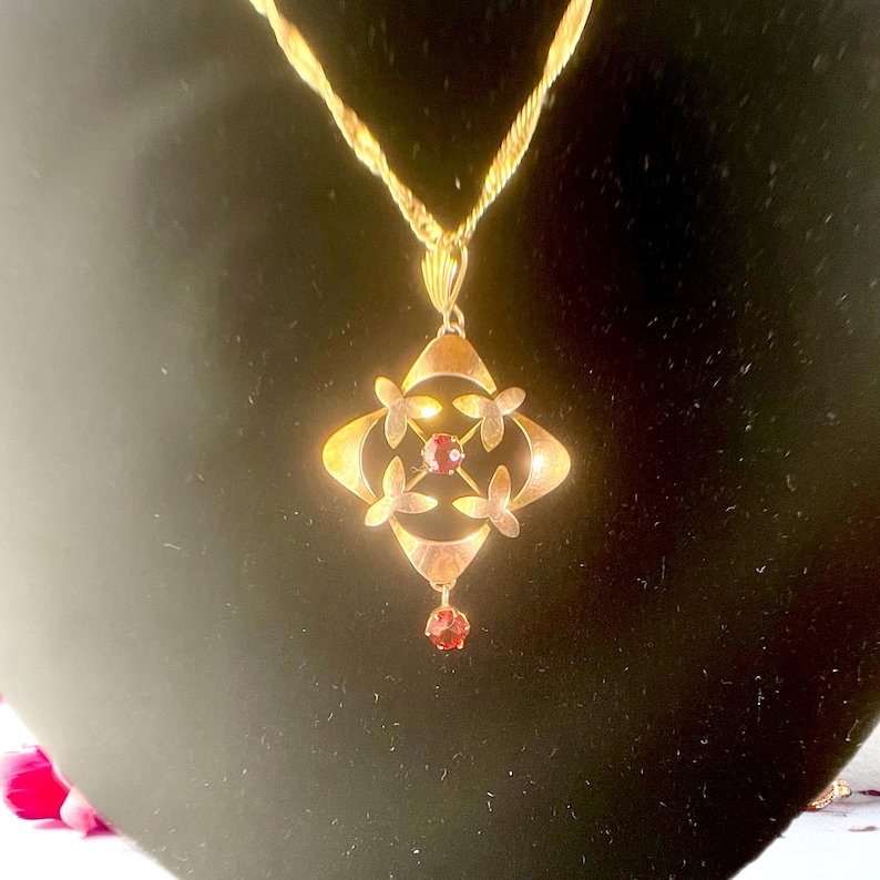 May include: A gold-toned pendant necklace with a decorative floral design. The pendant features a central red gemstone and four surrounding flower-shaped accents. A smaller red gemstone hangs below the pendant. The necklace chain is also gold-toned.