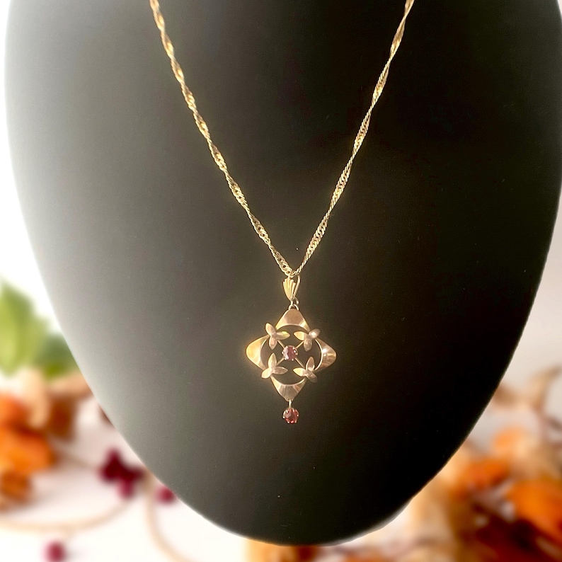 May include: A gold-colored pendant necklace with a delicate chain. The pendant features a four-leaf clover design with small red gemstones at the center and a single dangling gemstone. The necklace is displayed on a black mannequin.
