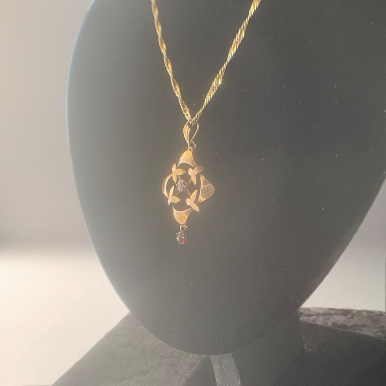 May include: A gold-colored pendant necklace with a floral design. The pendant features a four-petal flower shape with a small, round, red gemstone at the center and a teardrop-shaped red gemstone at the bottom. The chain is a twisted rope style.