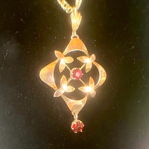 May include: Gold-toned pendant necklace with a floral design. The pendant features a central red gemstone surrounded by four stylized flower shapes. A smaller red gemstone hangs below. The necklace is on a gold chain.
