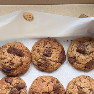 May include: Six chocolate chip cookies in a cardboard box. The cookies are a golden brown colour with large chunks of dark chocolate. A sticker on the box reads "handmade with love."