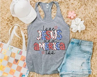 May include: A gray tank top with the words "Love Jesus and America too" in red, white, and blue. The image also includes a white baseball cap, denim shorts, a tote bag, a scrunchie, and a flower.