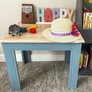 Könnte beinhalten: Ein hellblauer Holztisch mit einer weiß getünchten Tischplatte. Auf dem Tisch liegt ein Strohhut mit einem violetten Band und einer rosa Blume. Der Tisch steht auf einem grauen Teppich. Das Wort "HAPPY" ist in bunten Buchstaben hinter dem Tisch geschrieben.
