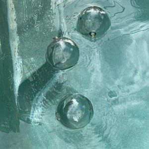May include: Three clear glass orbs float in a turquoise blue pool of water. The orbs are smooth and round, reflecting the light from above.