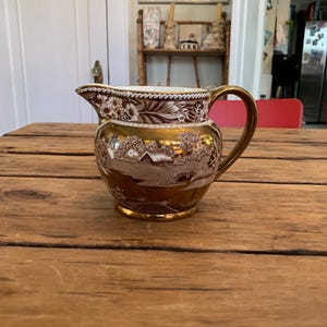May include: A ceramic creamer pitcher featuring a metallic gold finish and a brown transferware landscape scene. The pitcher has a curved handle, a pouring spout, and a decorative floral border pattern around the upper rim and neck area.