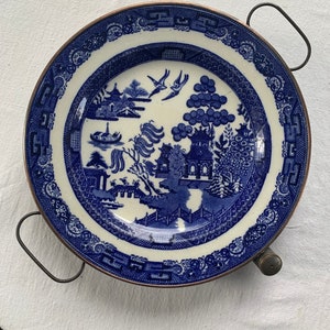 May include: A blue and white porcelain plate with a willow pattern design, set in a copper-colored metal frame with two handles.