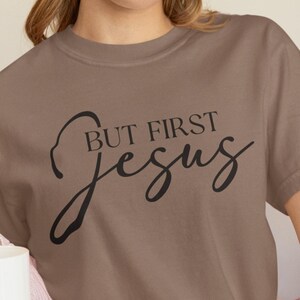 May include: A brown t-shirt with the text "BUT FIRST Jesus" in black script. The word "EXPRESSO" is in a black rectangle at the bottom of the image. The person is holding a white mug and wearing a pink sweater.