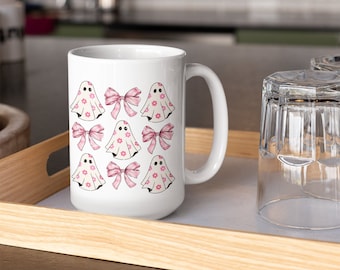 Puede incluir: Taza de cerámica blanca con un diseño de fantasmas rosa y blanco. Los fantasmas llevan lazos rosas y tienen pequeñas flores rosas sobre ellos.
