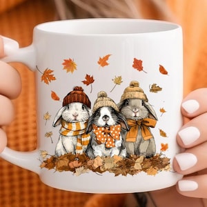 May include: White ceramic mug featuring three cartoon bunnies wearing hats and scarves, surrounded by autumn leaves. The bunnies have different colored fur and wear orange, yellow, and brown accessories. A cozy fall-themed design.