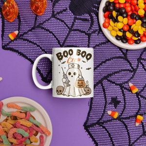 Op de afbeelding: Een witte keramische mok met een zwart-witte illustratie van een spook dat een verpleegstersmuts draagt en een spuitje vasthoudt. De mok heeft de tekst "Boo Boo Crew" in zwarte letters. De mok staat op een paarse achtergrond met een zwart spinnenwebpatroon en ma&iuml;s snoepjes.