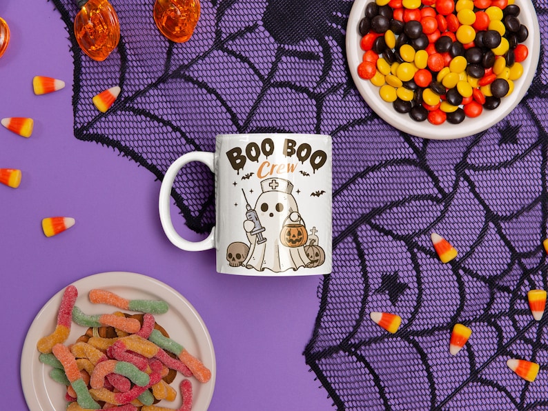 Op de afbeelding: Een witte keramische mok met een zwart-witte illustratie van een spook dat een verpleegstersmuts draagt en een spuitje vasthoudt. De mok heeft de tekst "Boo Boo Crew" in zwarte letters. De mok staat op een paarse achtergrond met een zwart spinnenwebpatroon en ma&iuml;s snoepjes.