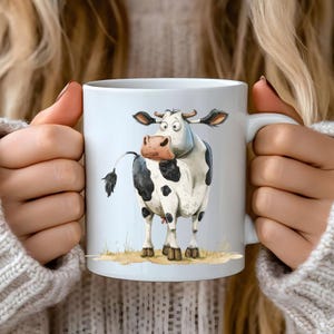 May include: White ceramic mug with a cartoon illustration of a black and white cow with big eyes. The cow is looking to the left of the mug.