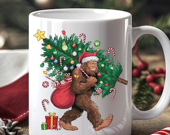 Puede incluir: Una taza de cerámica blanca con una ilustración colorida de una criatura Bigfoot marrón con gorro de Papá Noel, cargando un árbol de Navidad decorado y un saco rojo lleno de regalos, rodeado de bastones de caramelo y una estrella amarilla.