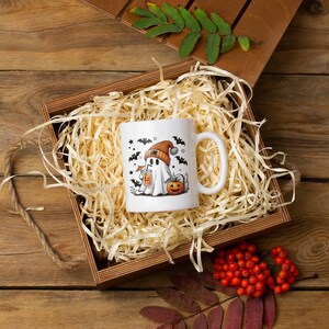 May include: A white ceramic mug with a Halloween-themed design featuring a ghost wearing a hat and holding a pumpkin. The mug is nestled in a brown wooden box filled with shredded paper.