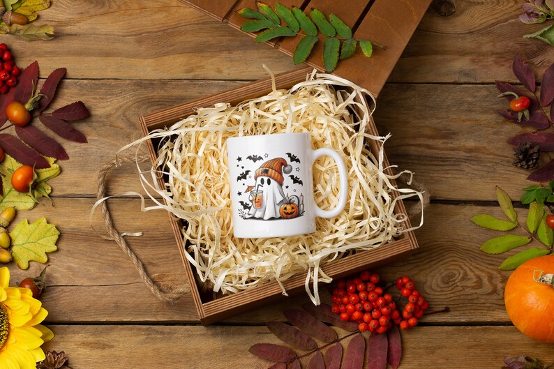 May include: A white ceramic mug with a Halloween-themed design featuring a ghost wearing a hat and holding a pumpkin. The mug is nestled in a brown wooden box filled with shredded paper.