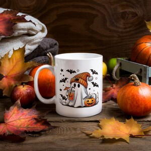 May include: White ceramic mug with a Halloween ghost design. The ghost is wearing a beanie and holding a drink. The mug is surrounded by fall leaves and pumpkins.