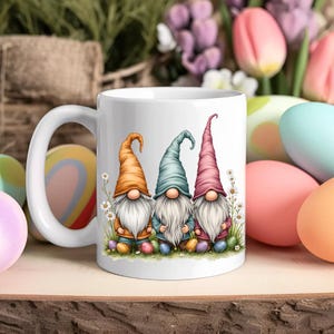 Könnte beinhalten: Weiße Keramiktasse mit einem Design aus drei Gnomen, die Ostereier halten. Die Gnomen tragen bunte Hüte und haben weiße Bärte. Die Tasse ist von pastellfarbenen Ostereiern umgeben.