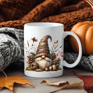 Peut inclure: Un mug en céramique blanche orné d'une illustration imprimée représentant un gnome avec un grand chapeau brun texturé, une longue barbe et un nez rond, entouré de pommes de pin, de feuillage séché et de petits glands.