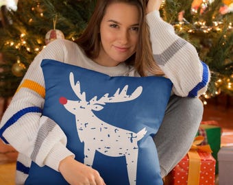 Puede incluir: Una almohada azul oscuro con un diseño de reno blanco y una nariz roja. La almohada está sentada en el regazo de una mujer frente a un árbol de Navidad.