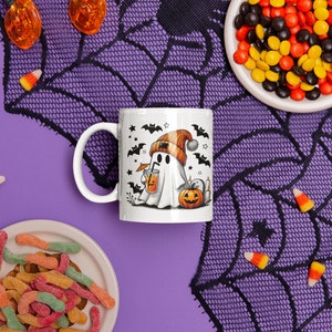 May include: White ceramic coffee mug with a black and white illustration of a ghost wearing a hat and holding a cup. The ghost is surrounded by bats and stars. The mug is on a purple surface with black and orange candy corn and a black spiderweb.