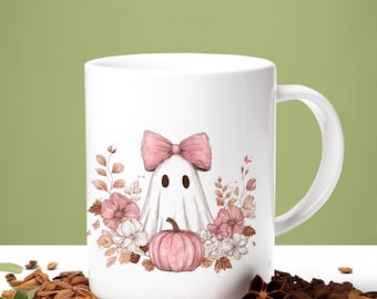 May include: A white ceramic mug featuring a graphic illustration of a white ghost wearing a large pink bow, positioned above a pink pumpkin and surrounded by a cluster of pink and brown flowers, leaves, and foliage on the front surface.