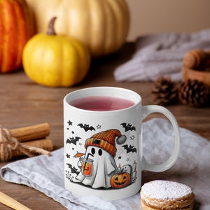 May include: White ceramic mug with a Halloween-themed design featuring a ghost wearing an orange beanie and holding a drink with an umbrella. The ghost is surrounded by bats, stars, and a pumpkin. The mug is filled with pink liquid and has a white handle.
