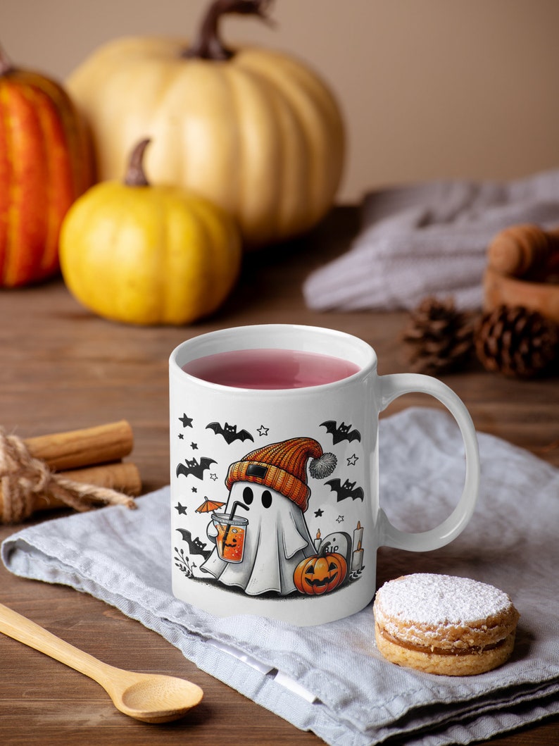 May include: White ceramic mug with a Halloween-themed design featuring a ghost wearing an orange beanie and holding a drink with an umbrella. The ghost is surrounded by bats, stars, and a pumpkin. The mug is filled with pink liquid and has a white handle.
