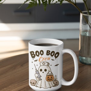 Op de afbeelding: Een witte keramische koffiemok met een zwarte handgreep. De mok heeft een cartoon-geest die een verpleegstersmuts draagt en een spuit en een pompoen vasthoudt. De tekst "Boo Boo Crew" staat op de mok gedrukt.