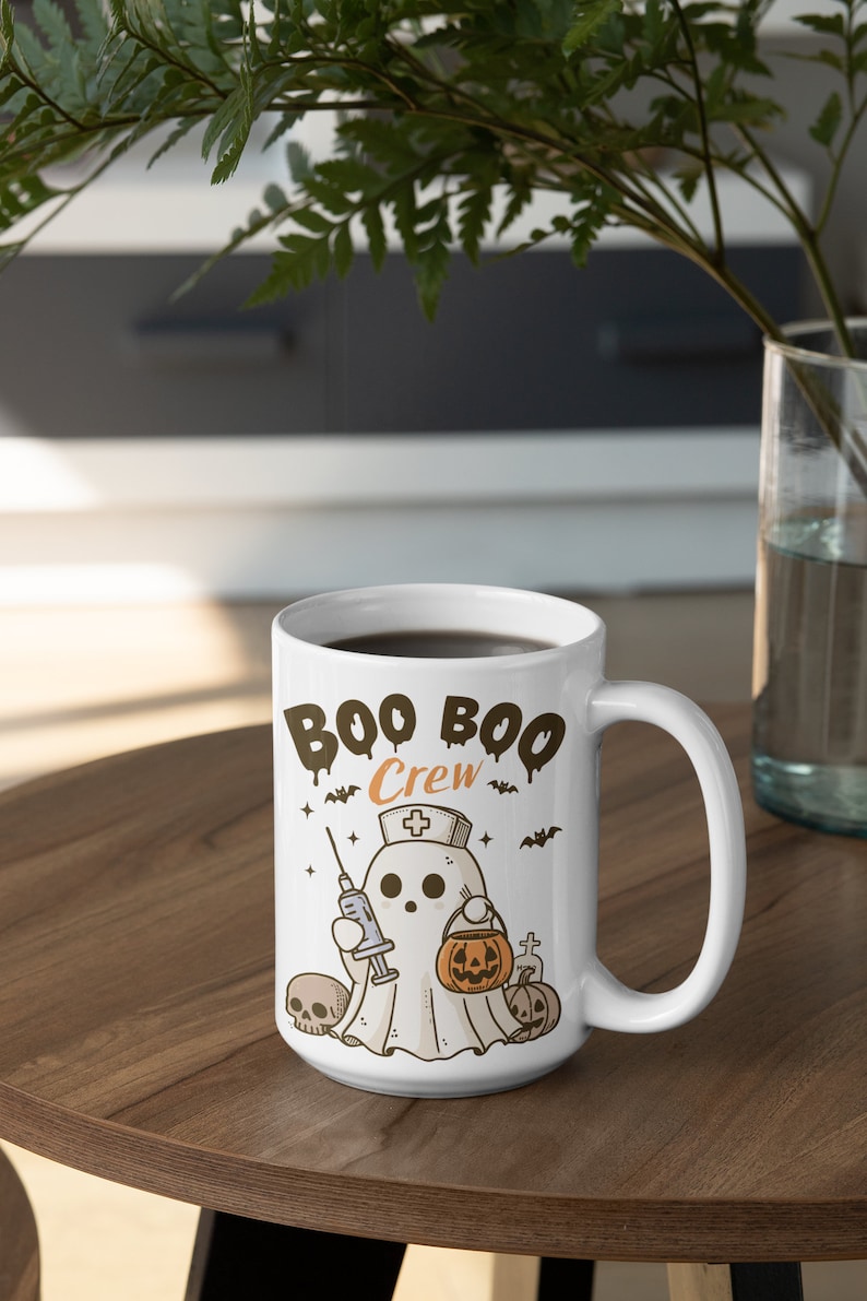 Op de afbeelding: Een witte keramische koffiemok met een zwarte handgreep. De mok heeft een cartoon-geest die een verpleegstersmuts draagt en een spuit en een pompoen vasthoudt. De tekst "Boo Boo Crew" staat op de mok gedrukt.