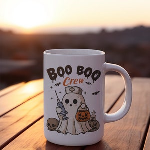 Op de afbeelding: Een witte keramische mok met een zwart-witte illustratie van een spook dat een verpleegstersmuts draagt en een spuit en een pompoen vasthoudt. De tekst "BOO BOO Crew" is in zwart op de mok gedrukt.