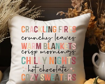 May include: A white throw pillow with the text "Crackling Fires, Crunchy Leaves, Warm Blankets, Crisp Mornings, Chilly Nights, Hot Chocolate, Cozy Sweaters" in colourful, stylised lettering.