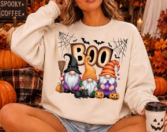 May include: A cream-colored crewneck sweatshirt featuring a graphic print of three gnomes wearing hats, holding a broom and a purple potion bottle, with a black cat, pumpkins, spiderwebs, bats, and the text BOO in a marquee light style.