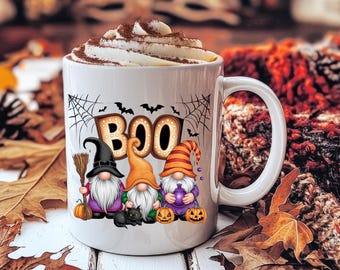 May include: A white ceramic mug filled with whipped cream topped with cocoa powder. The mug features a graphic of three gnomes, a black cat, pumpkins, spiderwebs, bats, and the text BOO in a marquee style font with light bulbs.
