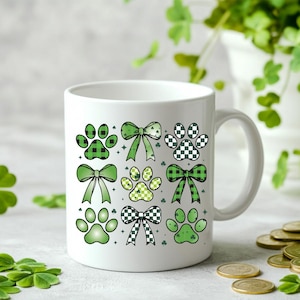 May include: White ceramic mug with a green and white paw print and bow design. The mug has a handle and is suitable for hot beverages.