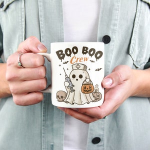 Op de afbeelding: Een witte keramische mok met een zwart-witte illustratie van een spook dat een verpleegstersmuts draagt en een spuit en een pompoen vasthoudt. De tekst "BOO BOO CREW" is in zwart op de mok gedrukt.
