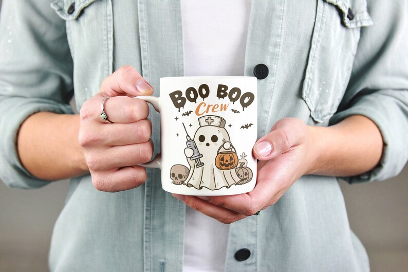 Op de afbeelding: Een witte keramische mok met een zwart-witte illustratie van een spook dat een verpleegstersmuts draagt en een spuit en een pompoen vasthoudt. De tekst "BOO BOO CREW" is in zwart op de mok gedrukt.