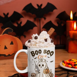 Op de afbeelding: Een witte keramische mok met een zwart-witte illustratie van een spook dat een verpleegstersmuts draagt en een spuit en een pompoen vasthoudt. De tekst "Boo Boo Crew" staat in zwarte, druppelende letters op de mok. De mok is gevuld met slagroom en een kaneelstokje.