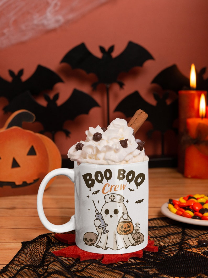 Op de afbeelding: Een witte keramische mok met een zwart-witte illustratie van een spook dat een verpleegstersmuts draagt en een spuit en een pompoen vasthoudt. De tekst "Boo Boo Crew" staat in zwarte, druppelende letters op de mok. De mok is gevuld met slagroom en een kaneelstokje.