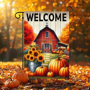 May include: A rectangular garden flag featuring the word WELCOME in black text above a red barn, a metal bucket filled with yellow sunflowers, and several orange pumpkins. The scene is set against a backdrop of autumn trees with orange foliage.