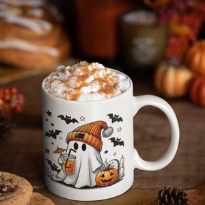 May include: White ceramic mug with a Halloween ghost design. The ghost is wearing an orange beanie and holding a glass of orange juice with an umbrella. The mug is filled with a creamy beverage topped with caramel and cinnamon.