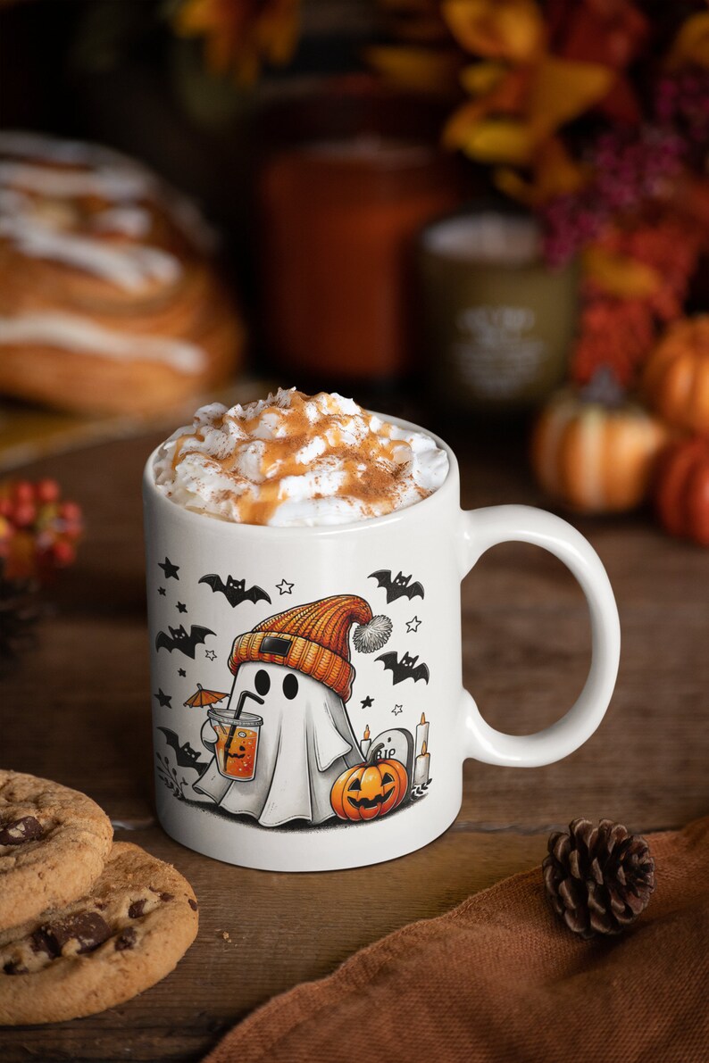May include: White ceramic mug with a Halloween ghost design. The ghost is wearing an orange beanie and holding a glass of orange juice with an umbrella. The mug is filled with a creamy beverage topped with caramel and cinnamon.