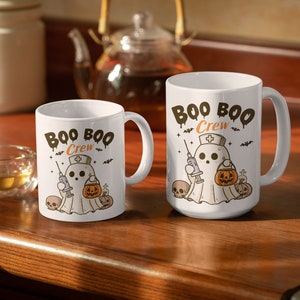 Op de afbeelding: Twee witte keramische mokken met een Halloween-thema. Beide mokken hebben een spook met een verpleegstersmuts die een spuit en een pompoen vasthoudt. De tekst op de mokken is "Boo Boo Crew".