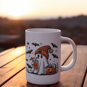May include: White ceramic mug with a Halloween-themed design. A ghost wearing a beanie hat holds a glass of orange juice. Bats, stars, and a pumpkin are also featured on the mug.