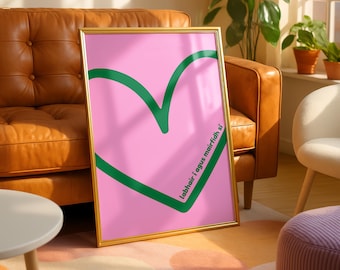 May include: A framed art print featuring a large green heart outline on a pink background. The artwork has a gold frame and includes text in the lower right corner. The print is displayed in a well-lit room with a leather sofa and plants.