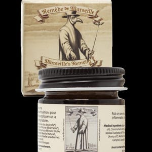 May include: A small jar of Marseille's Remedy with a black lid and a label featuring a plague doctor illustration. A matching box is behind the jar. The label text includes "Marseille's Remedy" and ingredient information.