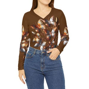 May include: A long-sleeved brown V-neck shirt featuring a butterfly design in shades of brown, orange, and gray. The butterflies are clustered together on the front and sleeves of the shirt. The model is wearing blue jeans.